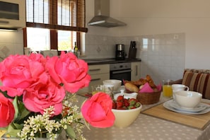 A kitchen with a dining table set for breakfast, including flowers, a bowl of fruit, bread, and cups.