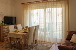 A dining area with a table set for four, a television on a stand, and a large window with sheer curtains.