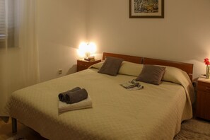 A neatly made bed with a quilted cover, two pillows, and a folded towel on the footboard. There is a bedside table with a lamp and a book. - Apartments Hrabar (Trogir)