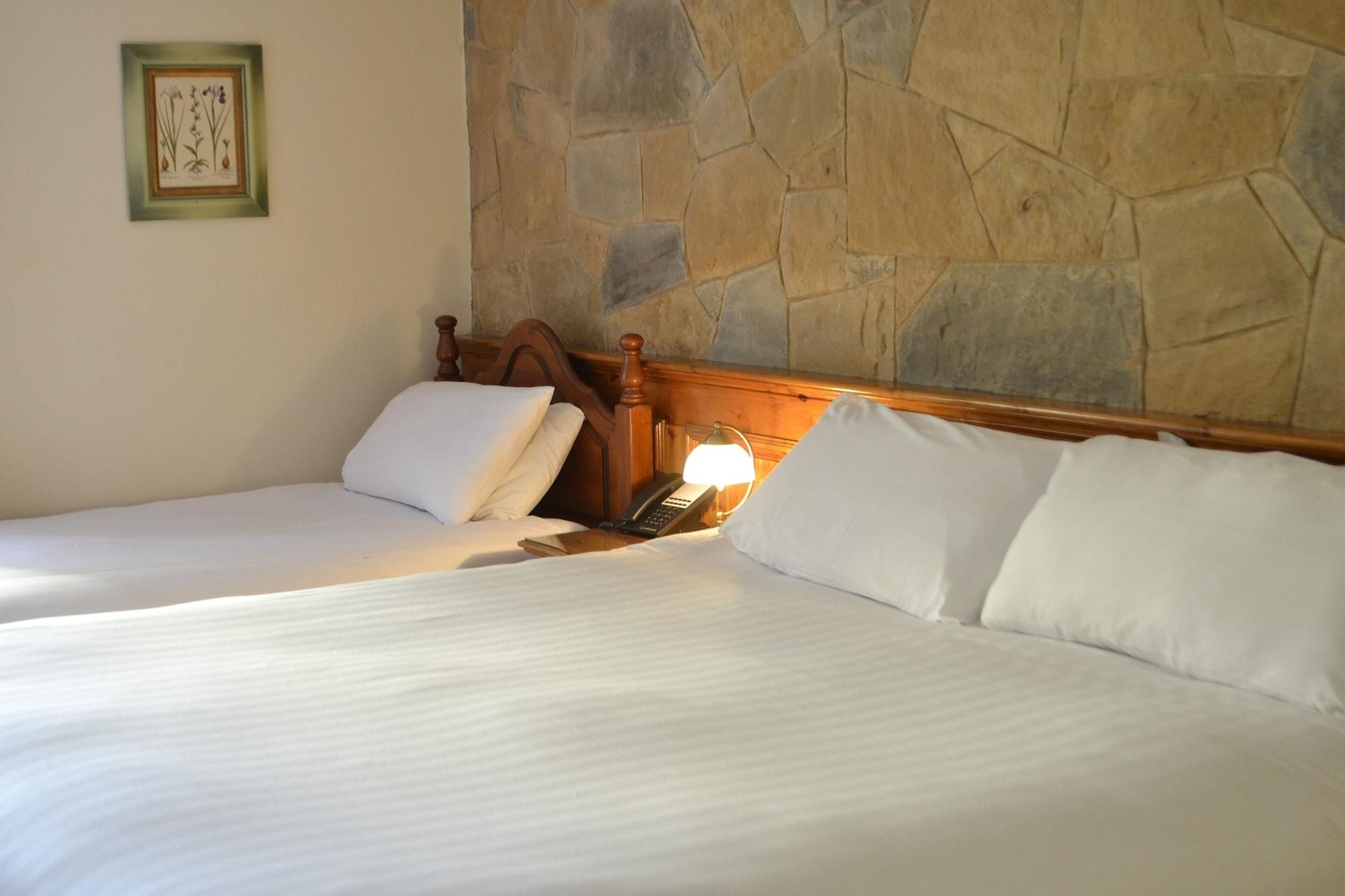 A double bed with white linens, a wooden headboard, a bedside table with a lamp, and a stone wall.