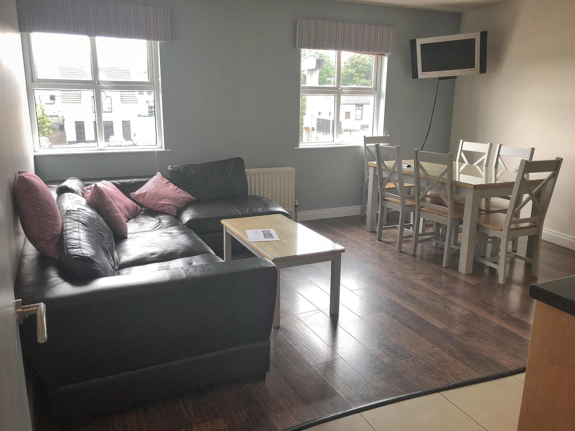 A living room with a black leather sofa, a wooden coffee table, a dining table with chairs, and a TV mounted on the wall.