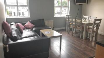 A living room with a black leather sofa, a wooden coffee table, a dining table with chairs, and a TV mounted on the wall.