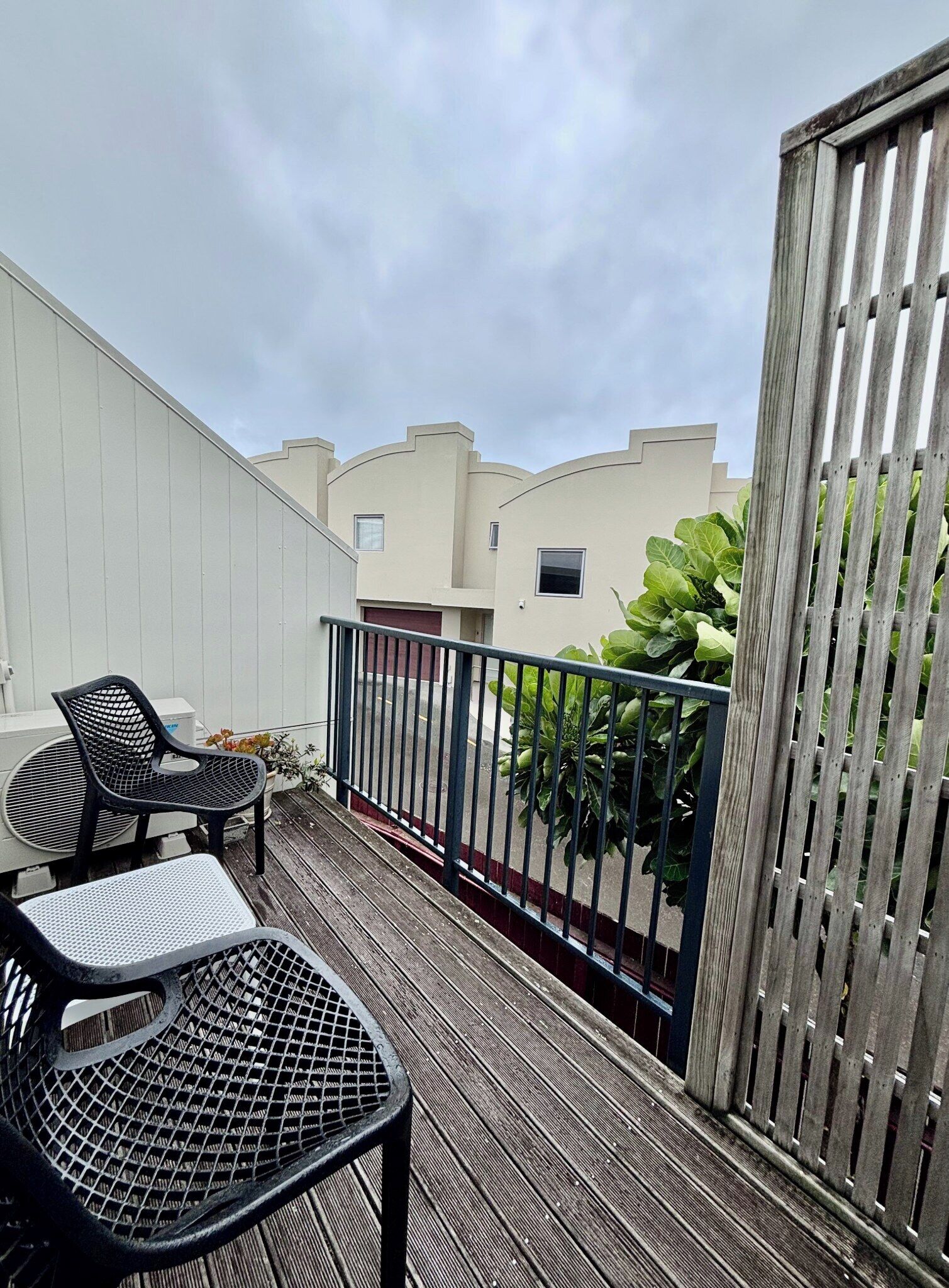 Un balcon avec deux chaises noires et une petite table, une clôture en bois et une vue sur les bâtiments voisins.