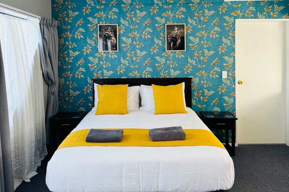 A neatly made bed with a white and yellow bedspread, two pillows, and two folded towels. The room has a blue floral wallpaper and a closed door.