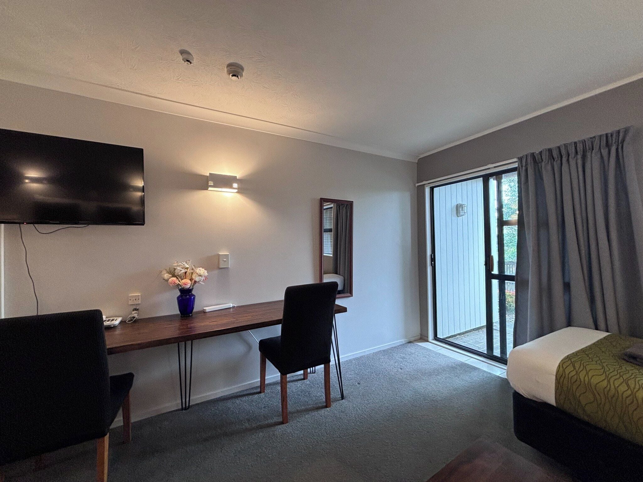 A hotel room with a bed, a desk, a chair, a TV, a vase with flowers, and a sliding glass door to the outside.