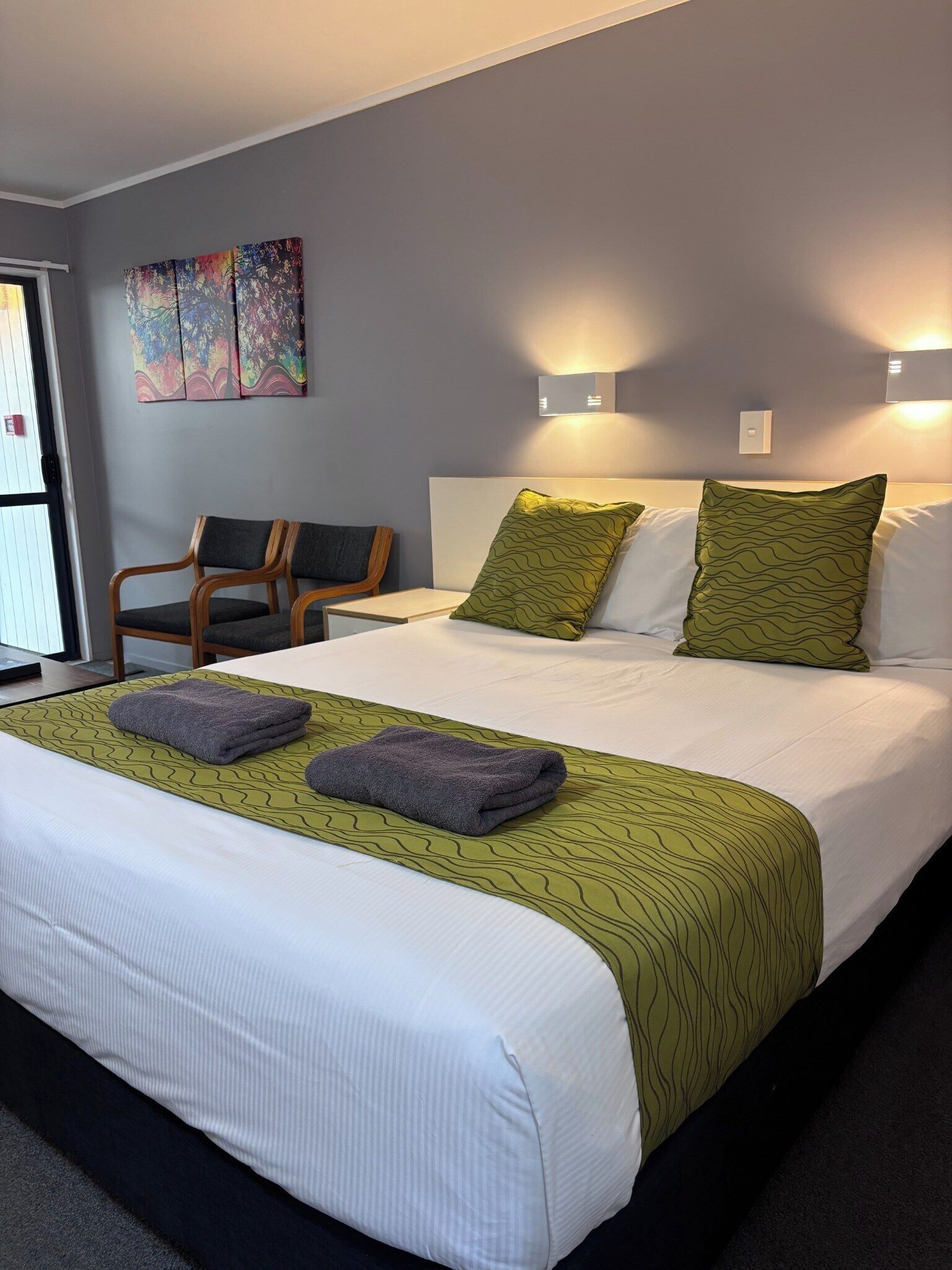 Une chambre d’hôtel avec un grand lit, deux coussins verts à motifs, une tête de lit blanche et une paire de chaises en bois.