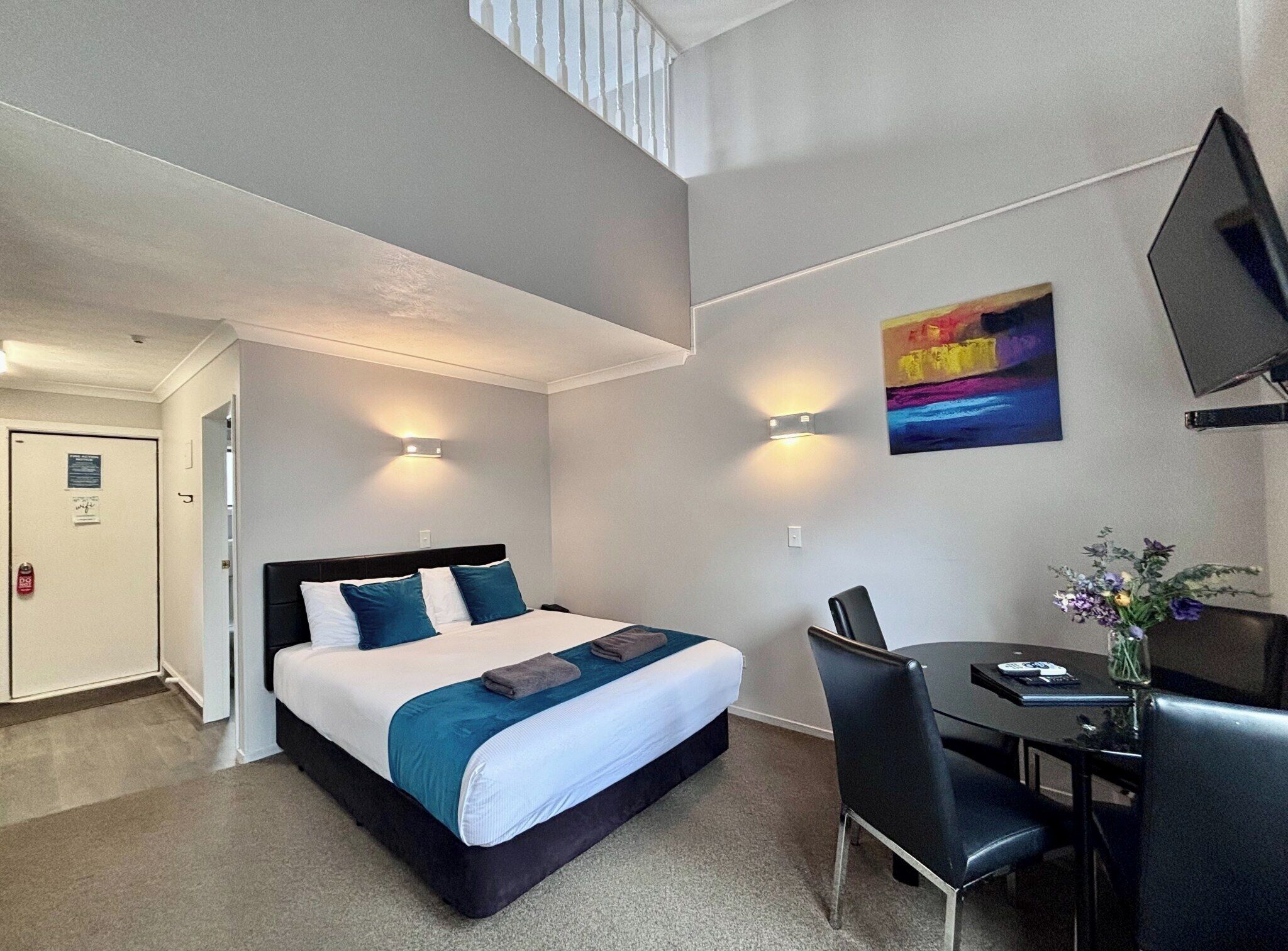 A hotel room with a bed, a desk, a chair, a TV, and a painting on the wall.