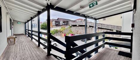 A covered outdoor balcony with a wooden floor, black railings, and a view of residential buildings.