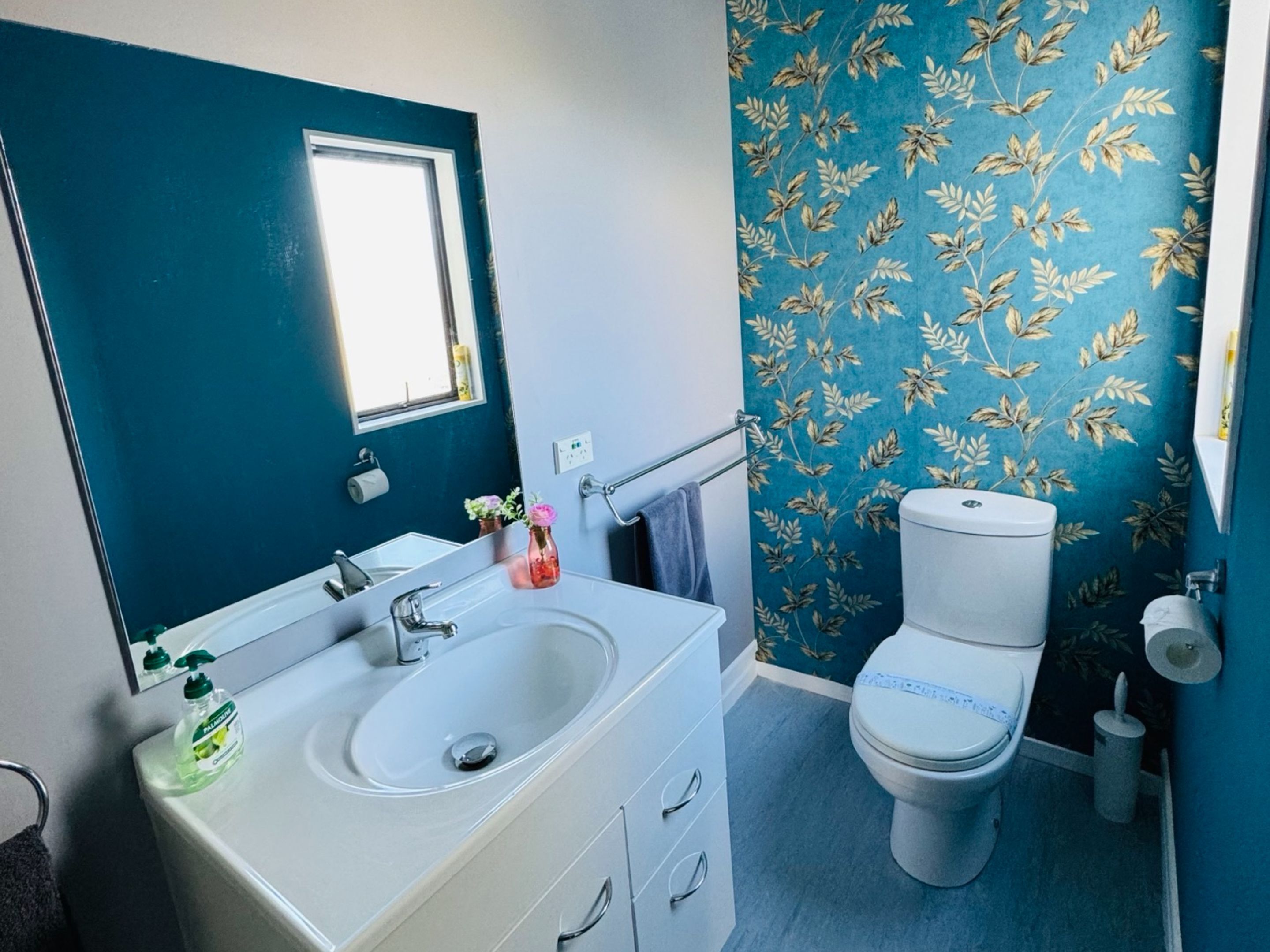 Casa de banho com lavatório branco, espelho, sanita e parede azul com padrão de folhagem.