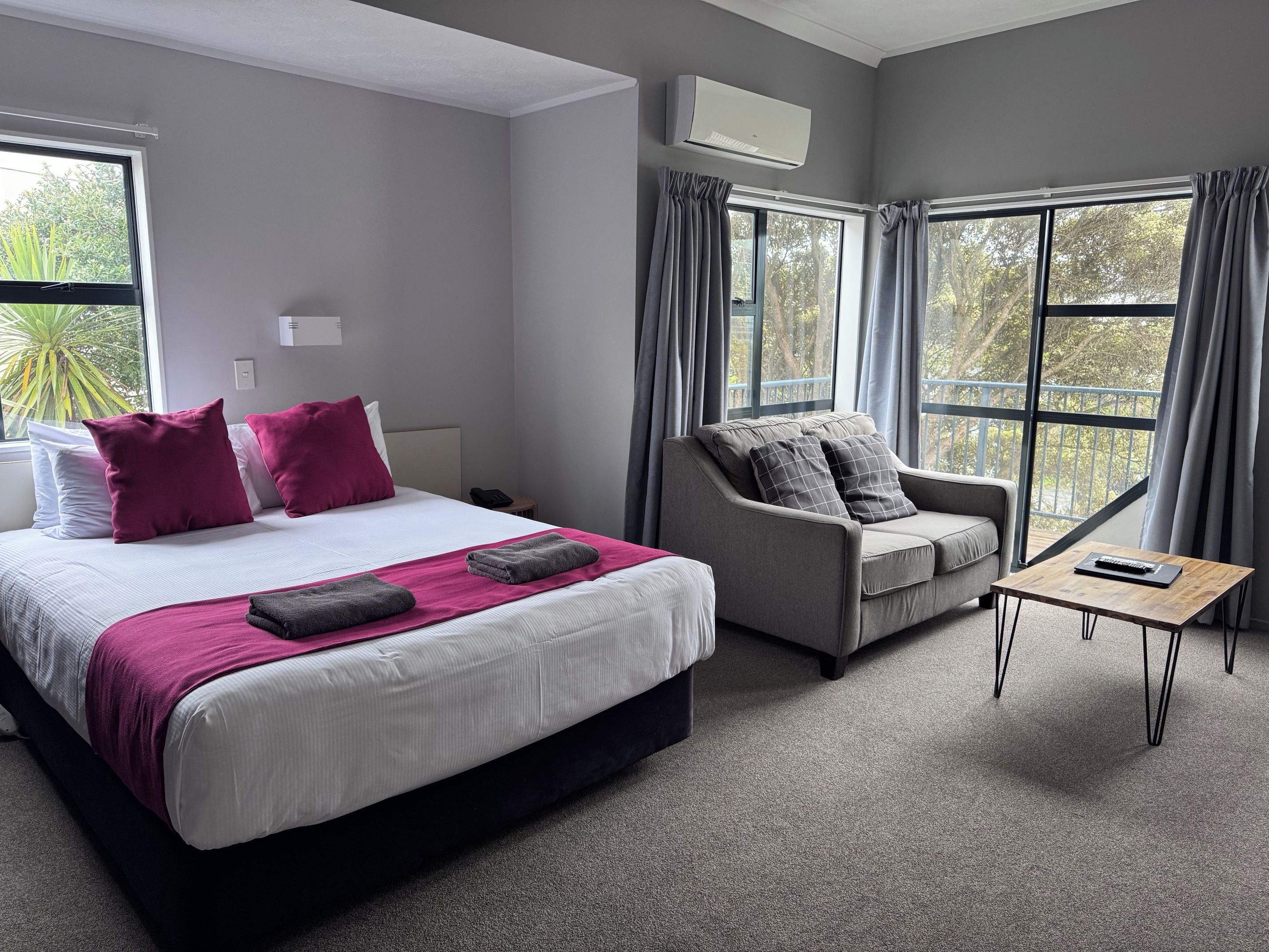 A hotel room with a bed, a grey sofa, a small table, and large windows.