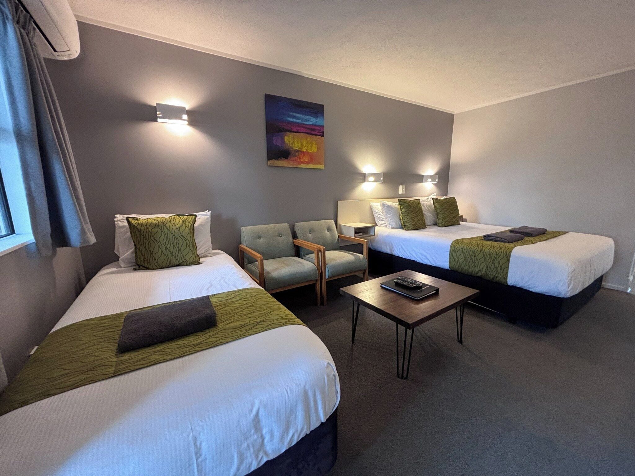 Habitación de hotel con dos camas, una silla, una mesa y un cuadro en la pared.