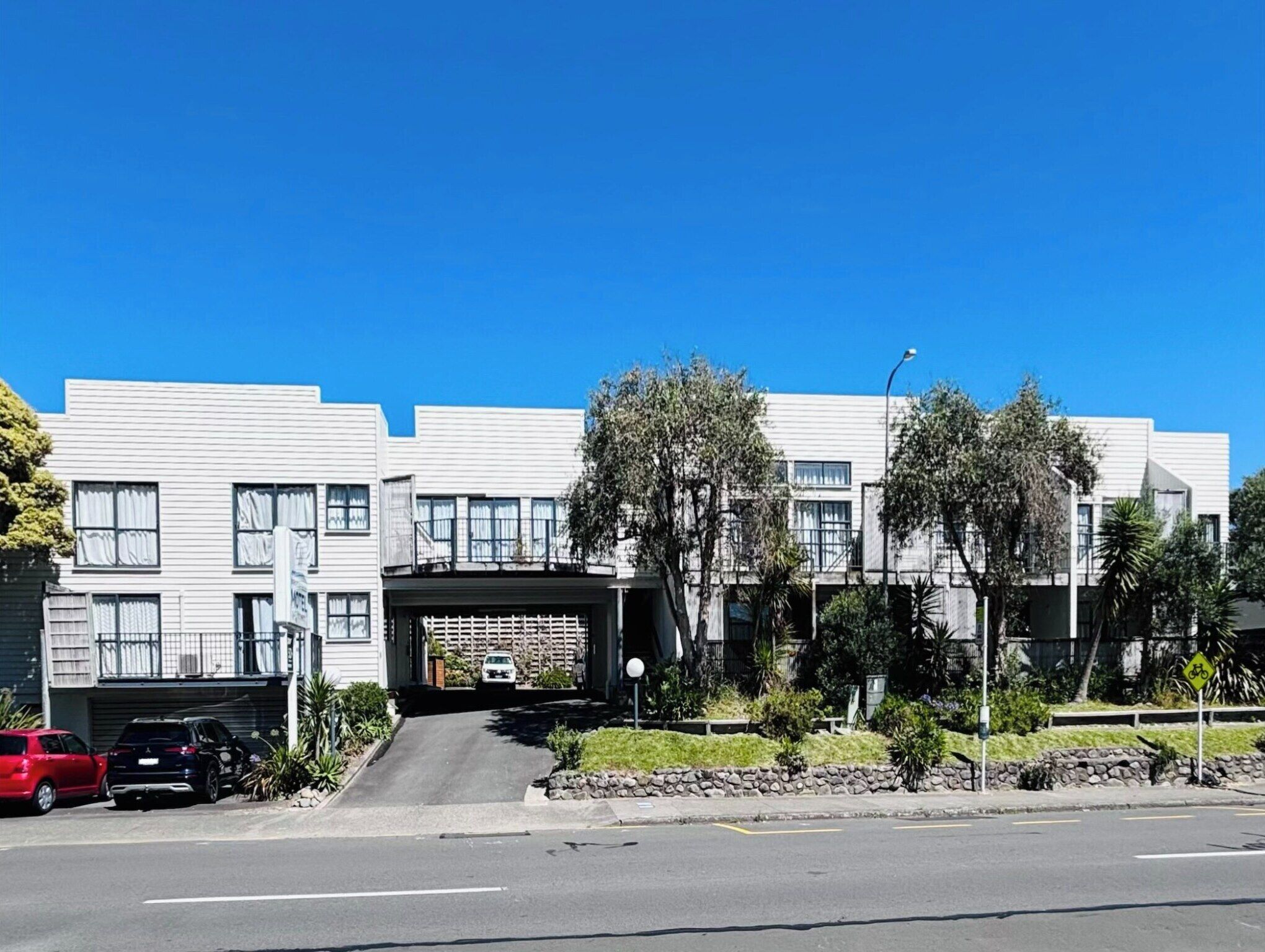 A modern multi-story residential building with a garage entrance, a clear blue sky, and a street with parked cars.