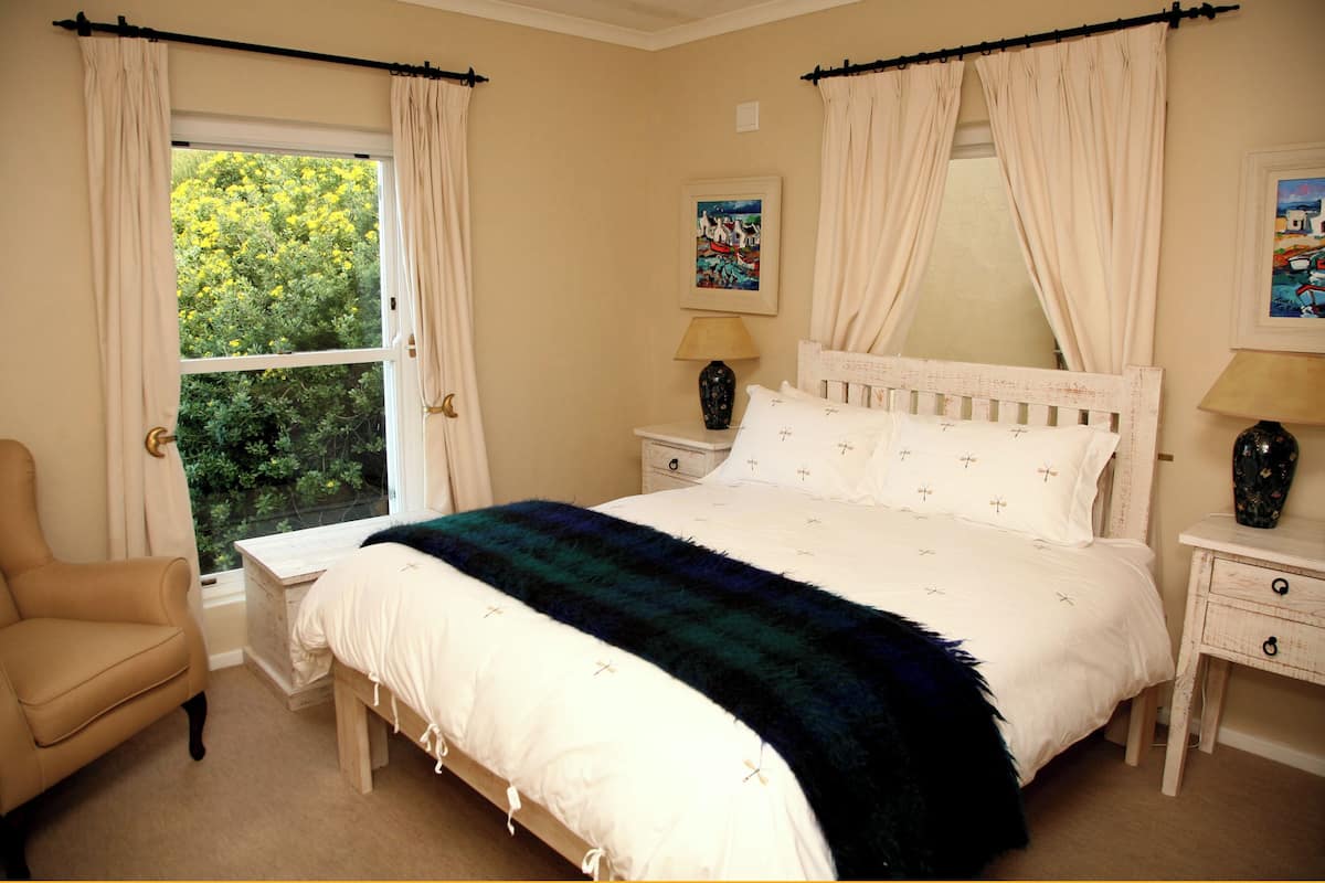 Ein Schlafzimmer mit Bett, Stuhl, Nachttisch, Lampe und einem Fenster mit Vorhängen.