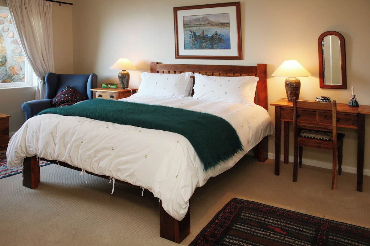 Cama bem arrumada com manta branca e verde, cabeceira de madeira, mesa de cabeceira com abajur, uma cadeira e um quadro na parede.