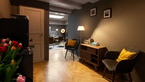 A modern living room with a wooden herringbone floor, a black sofa with a yellow pillow, a black armchair, and a side table with a lamp. A door leads to a dining area with a table and chairs. - Stanley Collection Hotel Hegra Amsterdam (Amsterdam)