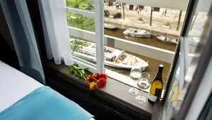 A hotel room with a bed, a window with a view of a canal and boats, a plate of food, a bottle of wine, and a bouquet of flowers. - Stanley Collection Hotel Hegra Amsterdam (Amsterdam)