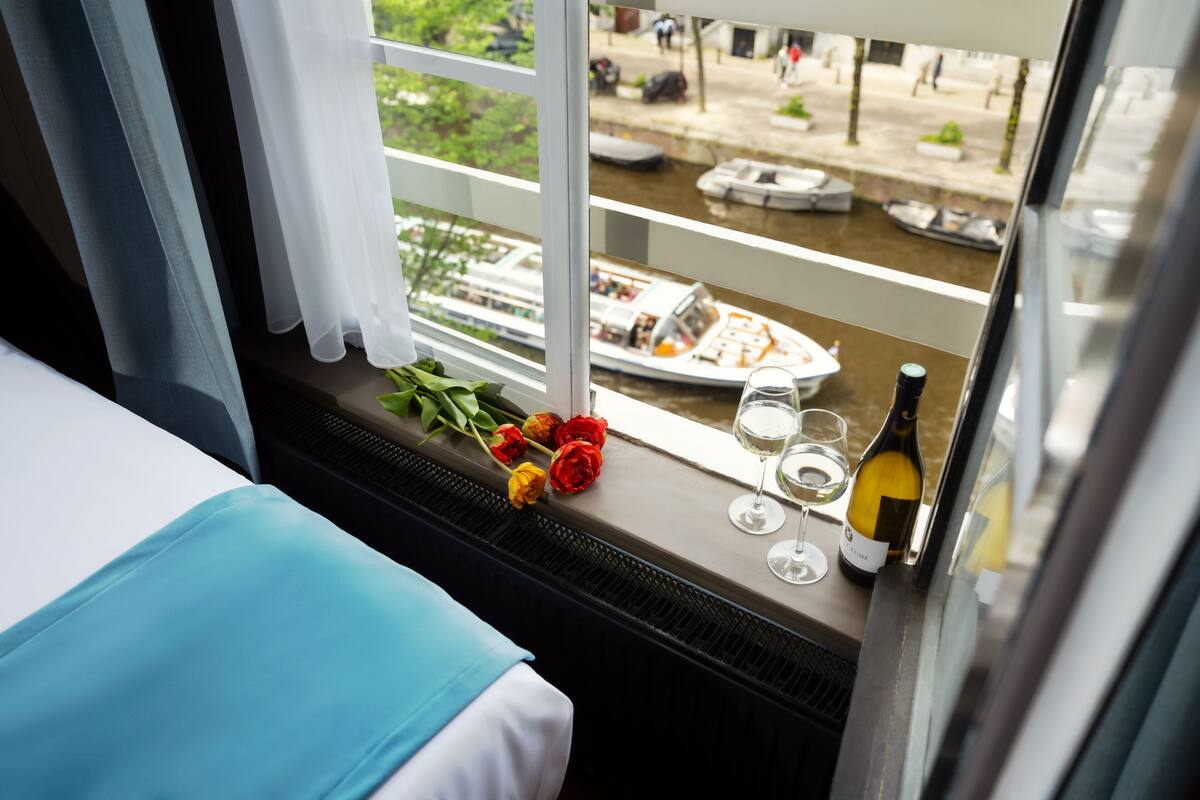 A hotel room with a bed, a window with a view of a canal and boats, a plate of food, a bottle of wine, and a bouquet of flowers.