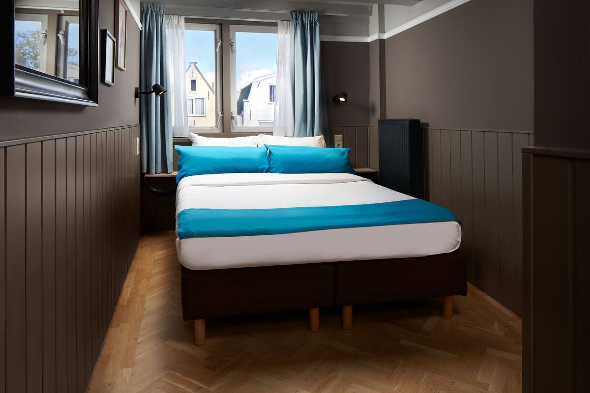 A bedroom with a large bed, wooden floor, a window with curtains, and a view of buildings outside.