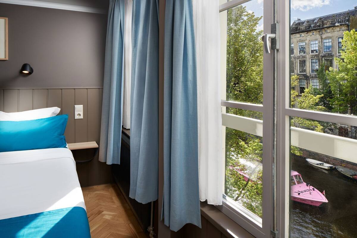 A hotel room with a bed, a window with curtains, a view of a canal with boats, and a building across the water.