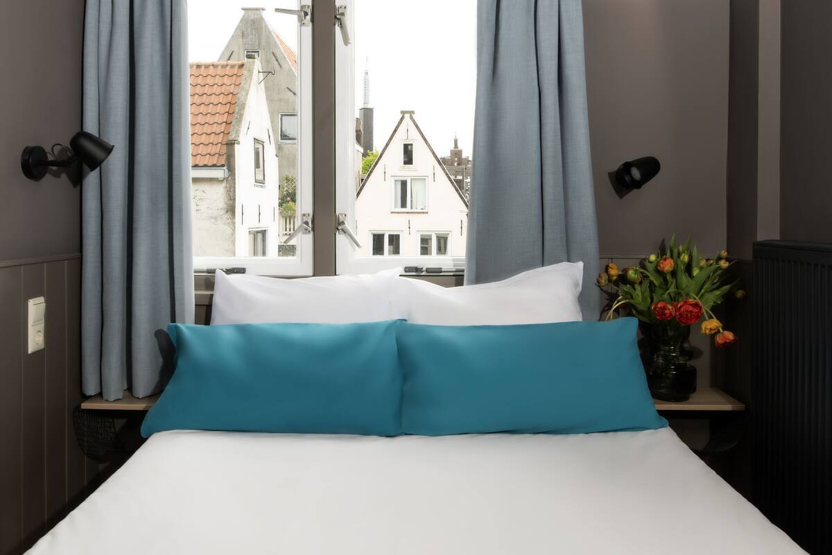 A hotel room with a bed, curtains, a vase with flowers, and a view of buildings outside.