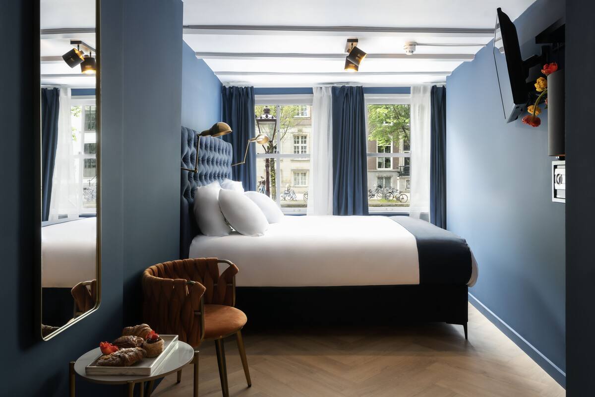 A hotel room with a large bed, a chair, a small table with pastries, and a window with curtains.