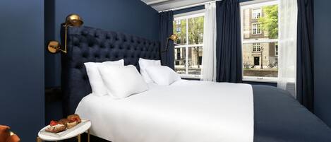 A bedroom with a large bed, a bedside table with pastries, and a view of buildings through the window.