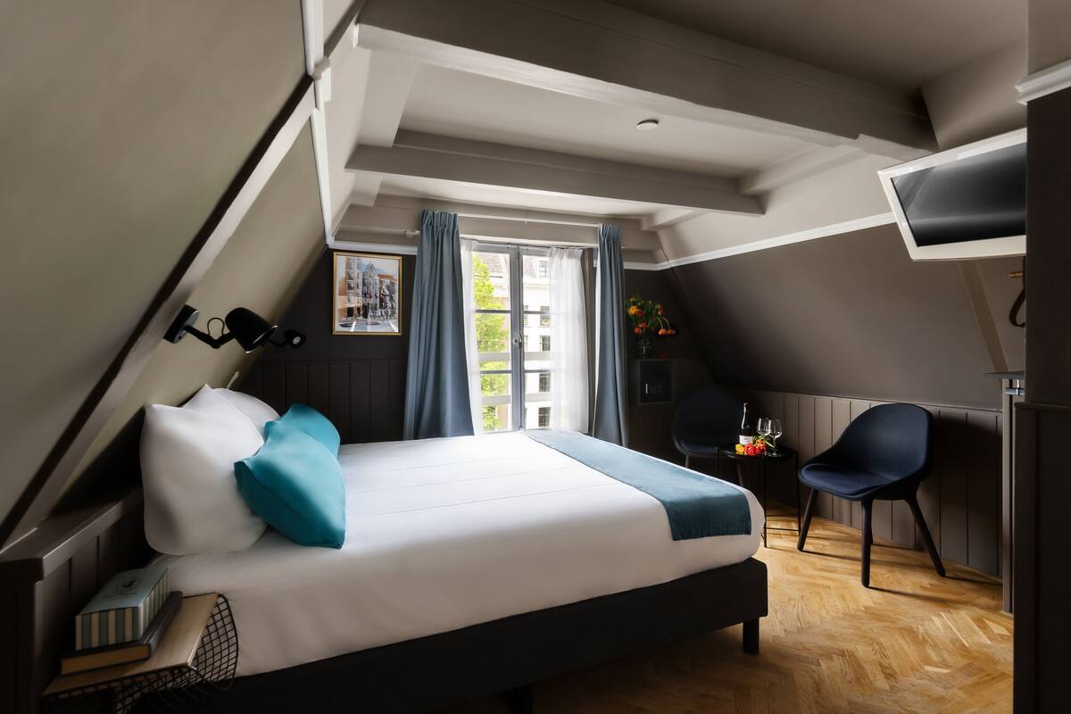A modern hotel room with a large bed, a desk with a chair, a window with curtains, and a view of buildings outside.