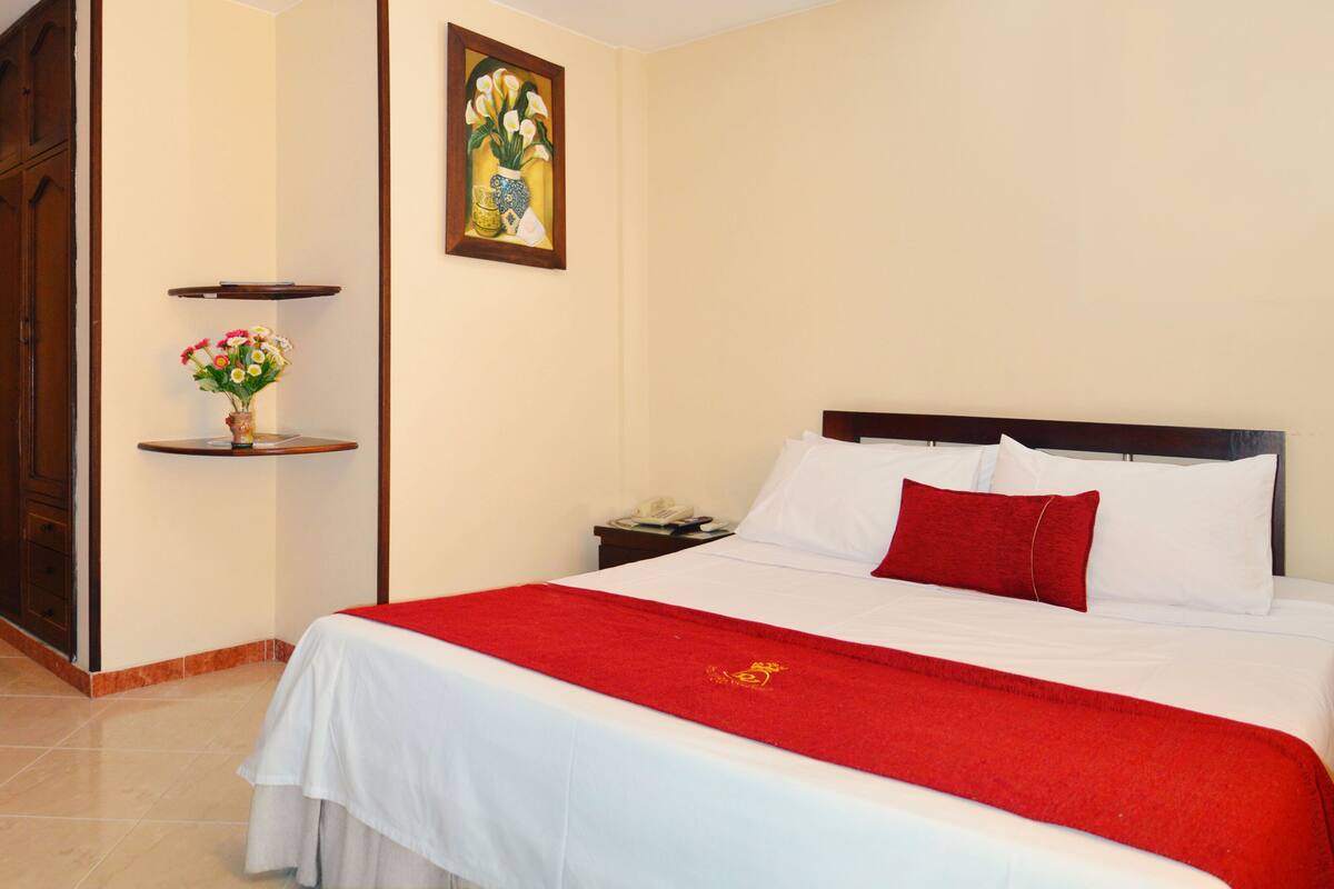 A hotel room with a bed, a red throw, a vase with flowers, and a painting on the wall.