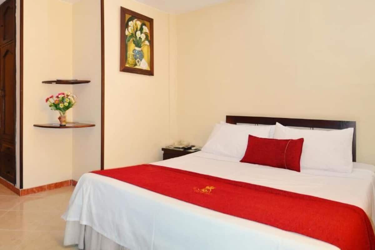 A hotel room with a bed, a nightstand, a red throw, a painting, and a wooden wardrobe.