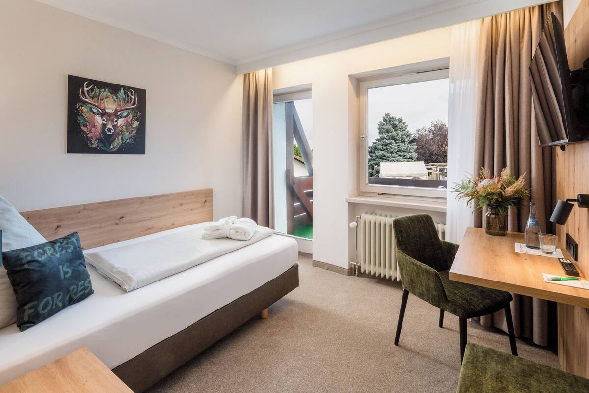 Une chambre d’hôtel moderne équipée d’un lit, d’un bureau, d’une chaise, d’une fenêtre avec des rideaux, d’un radiateur et d’un tableau accroché au mur.
