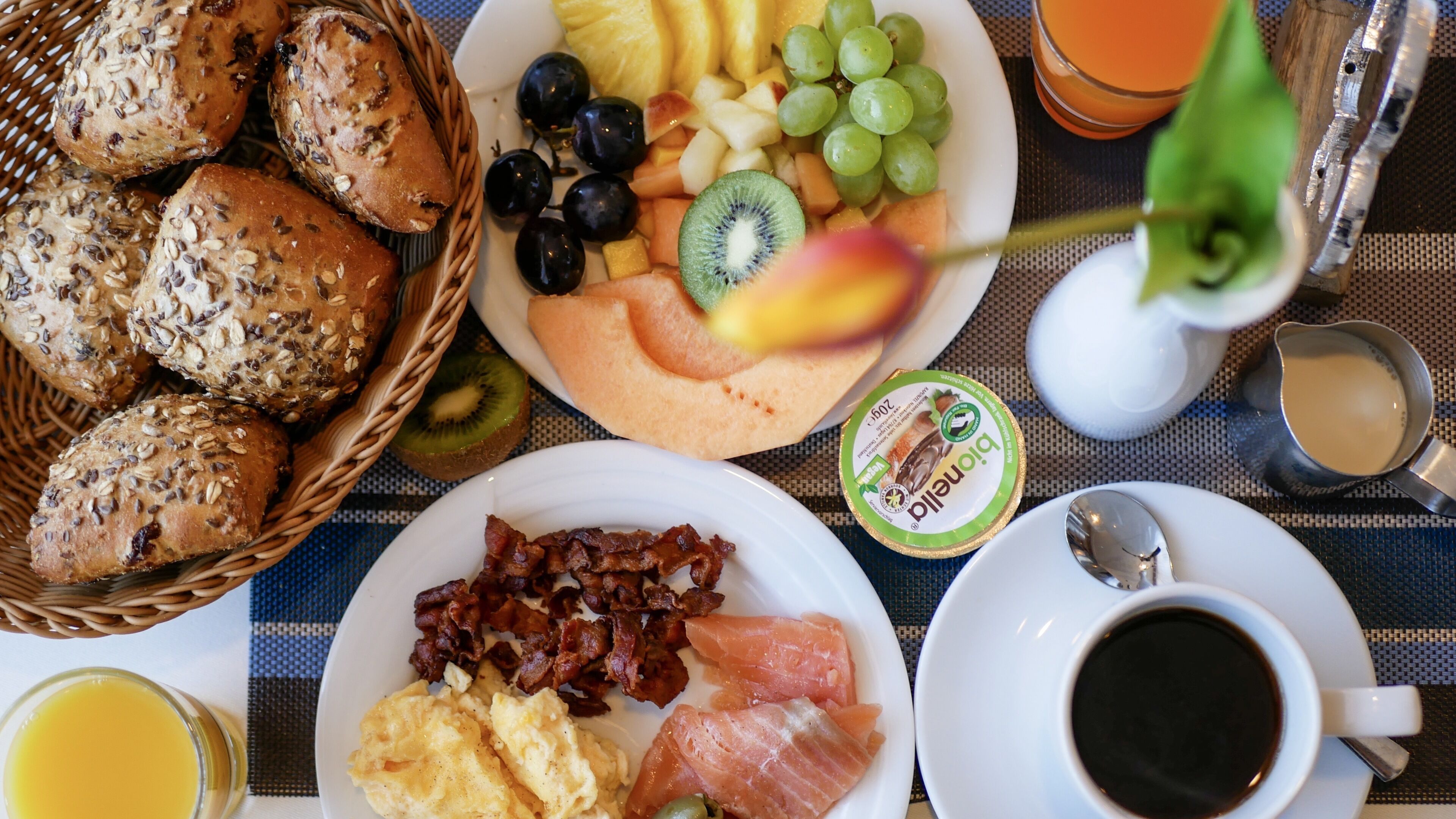 A breakfast table with bread, fruits, eggs, bacon, salmon, and coffee.