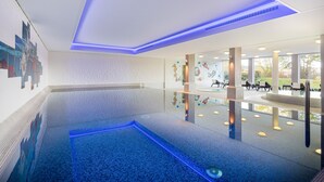 Une piscine intérieure moderne, dotée d’un bord illuminé de lumière bleue, de parois en verre transparent et offrant une vue sur un espace de détente extérieur.