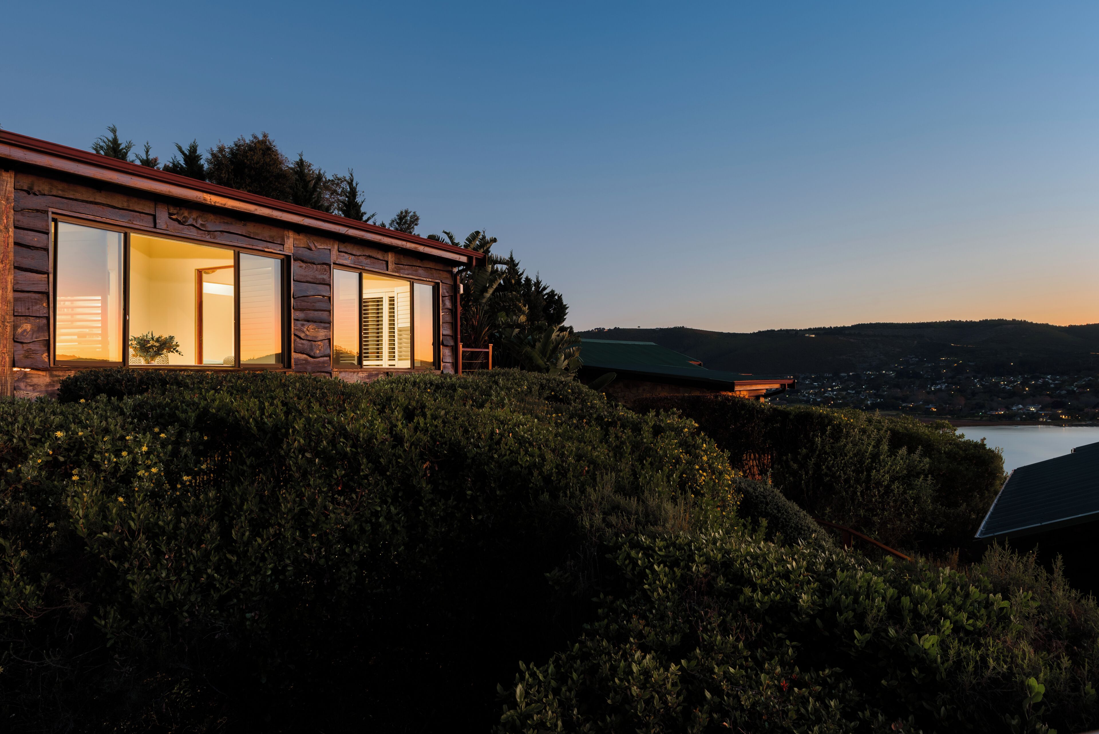A modern log cabin with large windows, a view of a lake, and a hillside landscape.