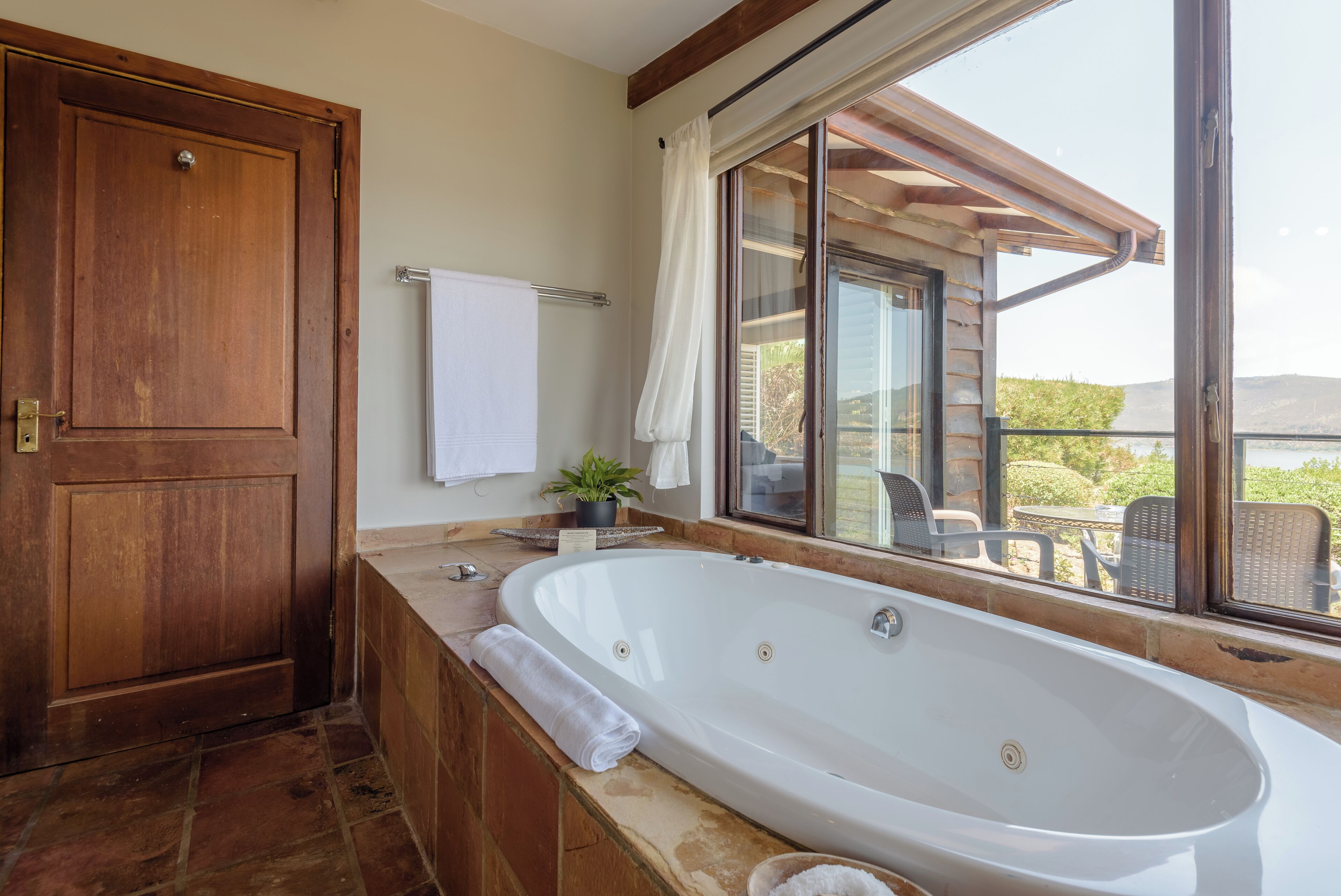 A bathroom with a large bathtub, a wooden door, a towel rack, and a window with a view of the outdoors.