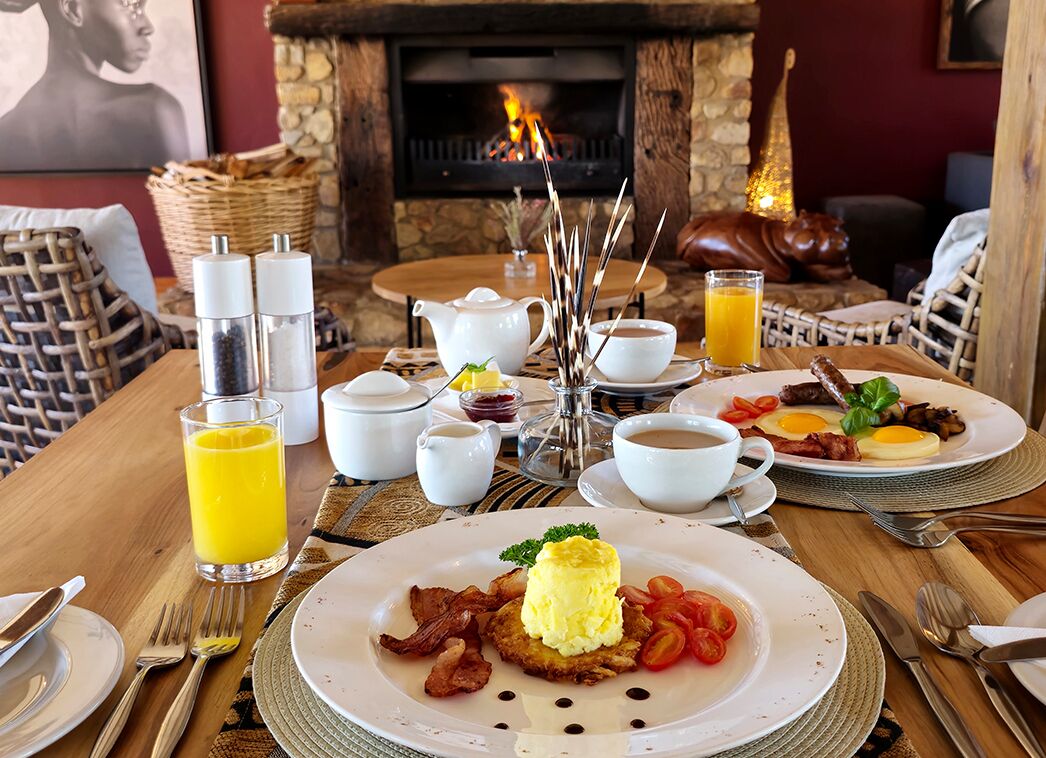 A breakfast setting with plates of eggs, bacon, and tomatoes, a glass of orange juice, and a fireplace in the background.