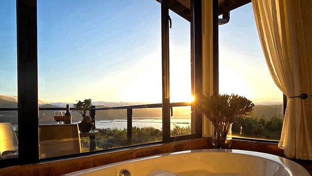 A large bathtub with a view of a sunset over a body of water, a potted plant, and a window with curtains. - Elephant Hide of Knysna Guest Lodge (Knysna)