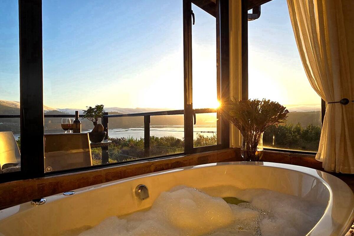 A large bathtub with a view of a sunset over a body of water, a potted plant, and a window with curtains.