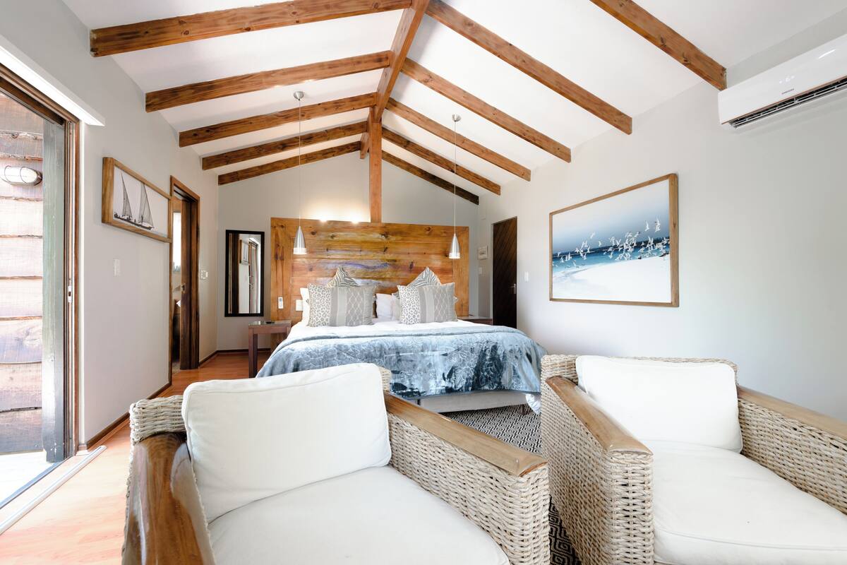 A bedroom with a bed, two wicker chairs, wooden beams, a large window, and a framed picture on the wall.