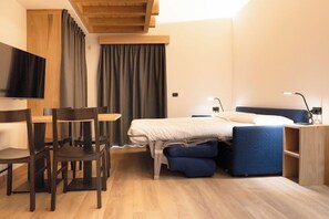 Quarto de hotel pequeno com cama, escrivaninha, cadeira, televisão e janela com cortinas.