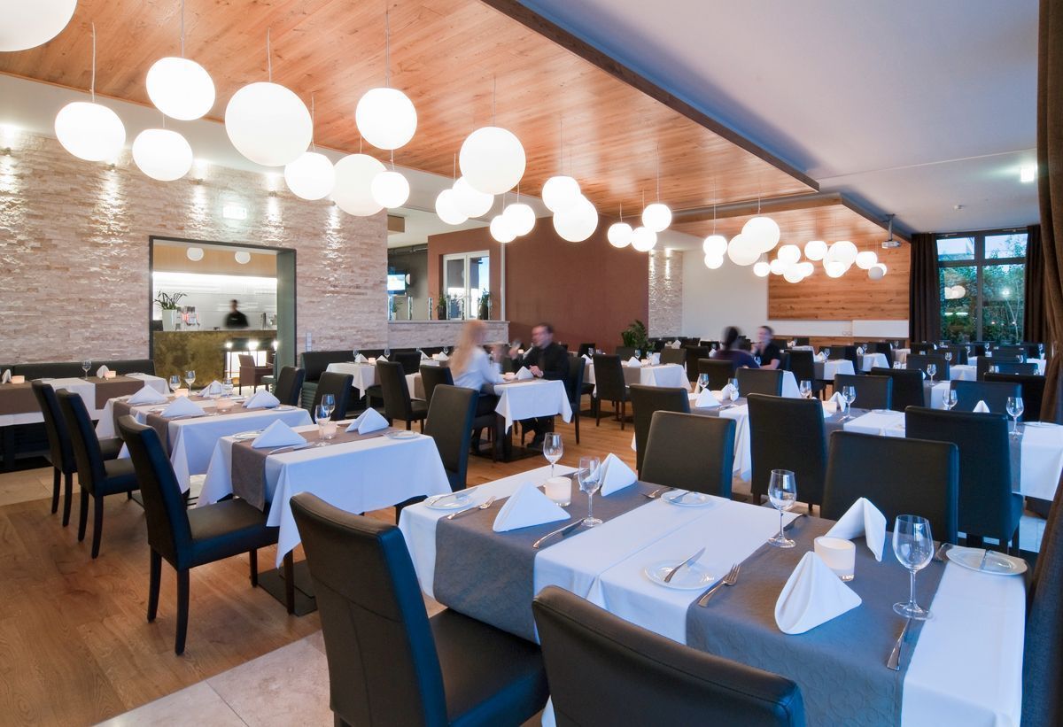 A restaurant with multiple tables set for dining, wooden ceiling, and hanging spherical lights.