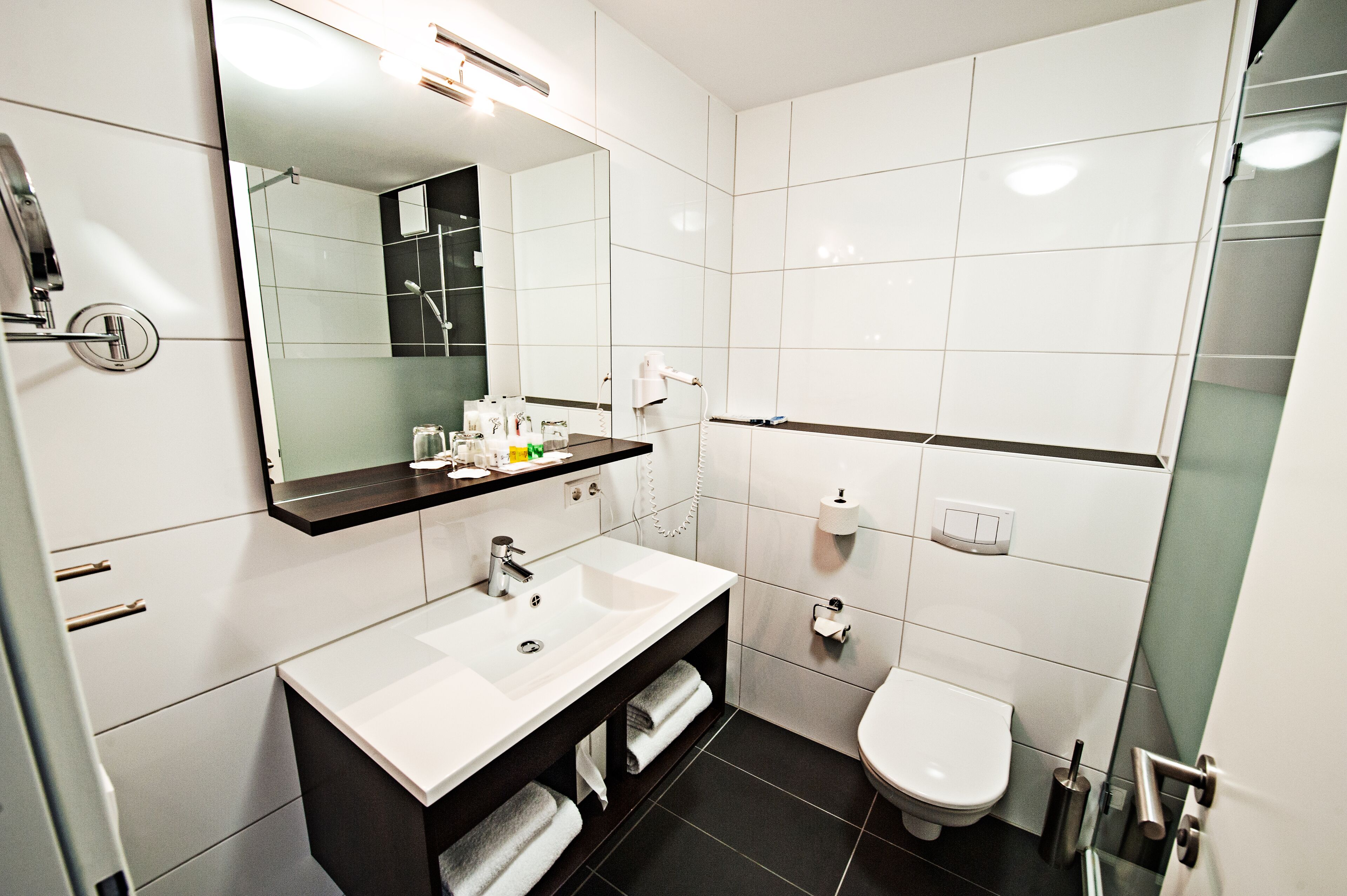 A modern bathroom with a white sink, dark floor tiles, a large mirror, and a toilet.
