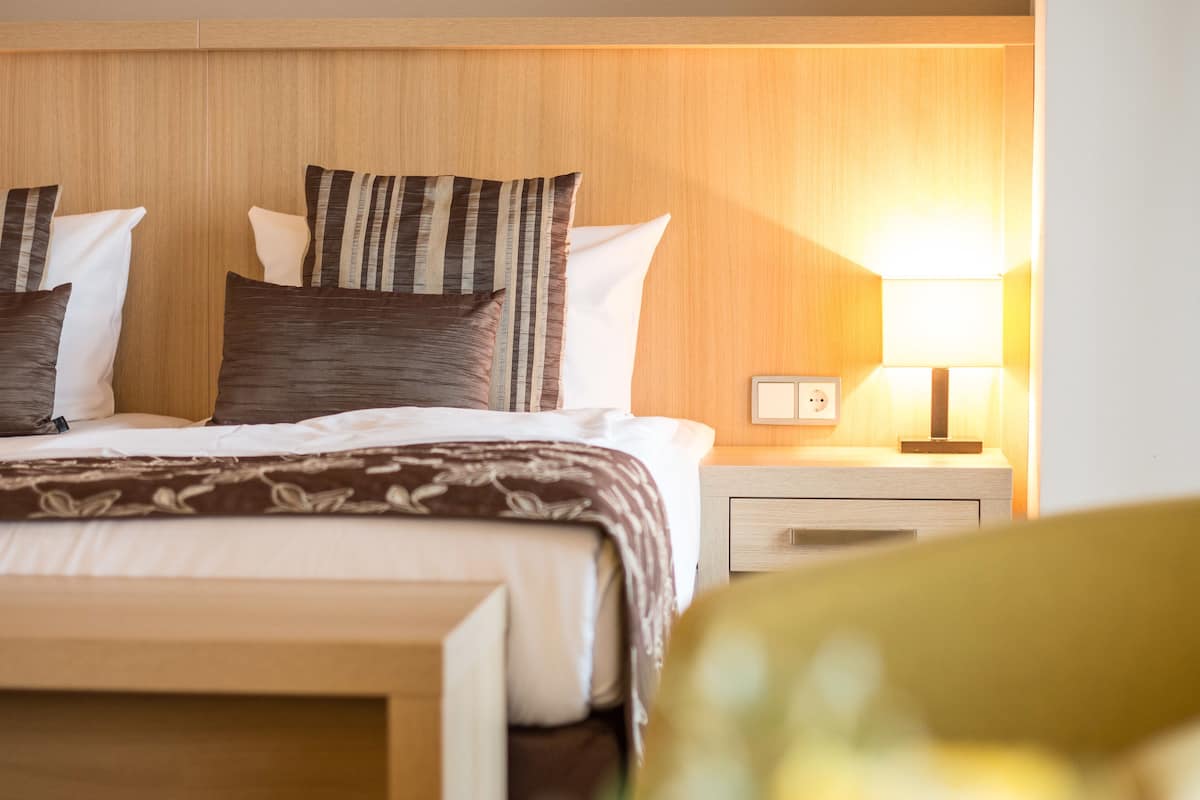 Een netjes opgemaakt bed met een beddeken met dessin en kussens, een nachtkastje met een lamp, en een houten hoofdbord.