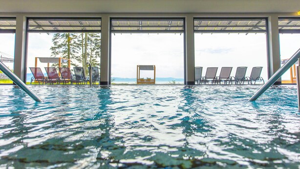 An indoor swimming pool with clear water, a wooden ceiling, and a view of the outdoors through large windows. - Bodensee Hotel Sonnenhof (Kressbronn)