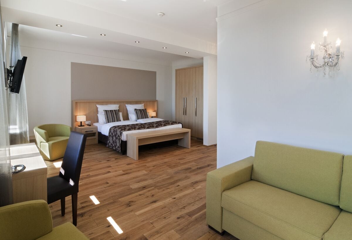 Een moderne hotelkamer met een houten vloer, een bed met een houten hoofdbord, een groene fauteuil, een bank, een bureau met een stoel en een aan de muur bevestigde televisie.