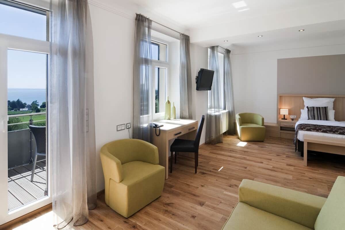 Een moderne hotelkamer met een houten vloer, een bed, een bureau met stoel, een bank en een aan de muur bevestigde televisie.