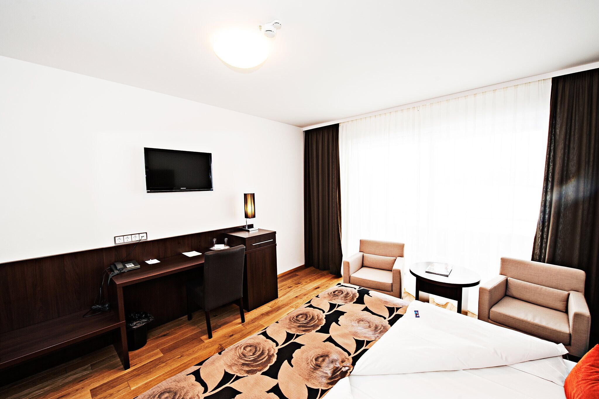 A hotel room with a bed, a desk, two chairs, a TV, and a window with curtains.