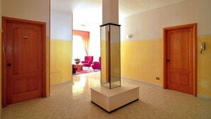 A hallway with two wooden doors, a central column, and a bright room in the background with a pink chair and a plant. - Albergo Paradiso (Monte Compatri)
