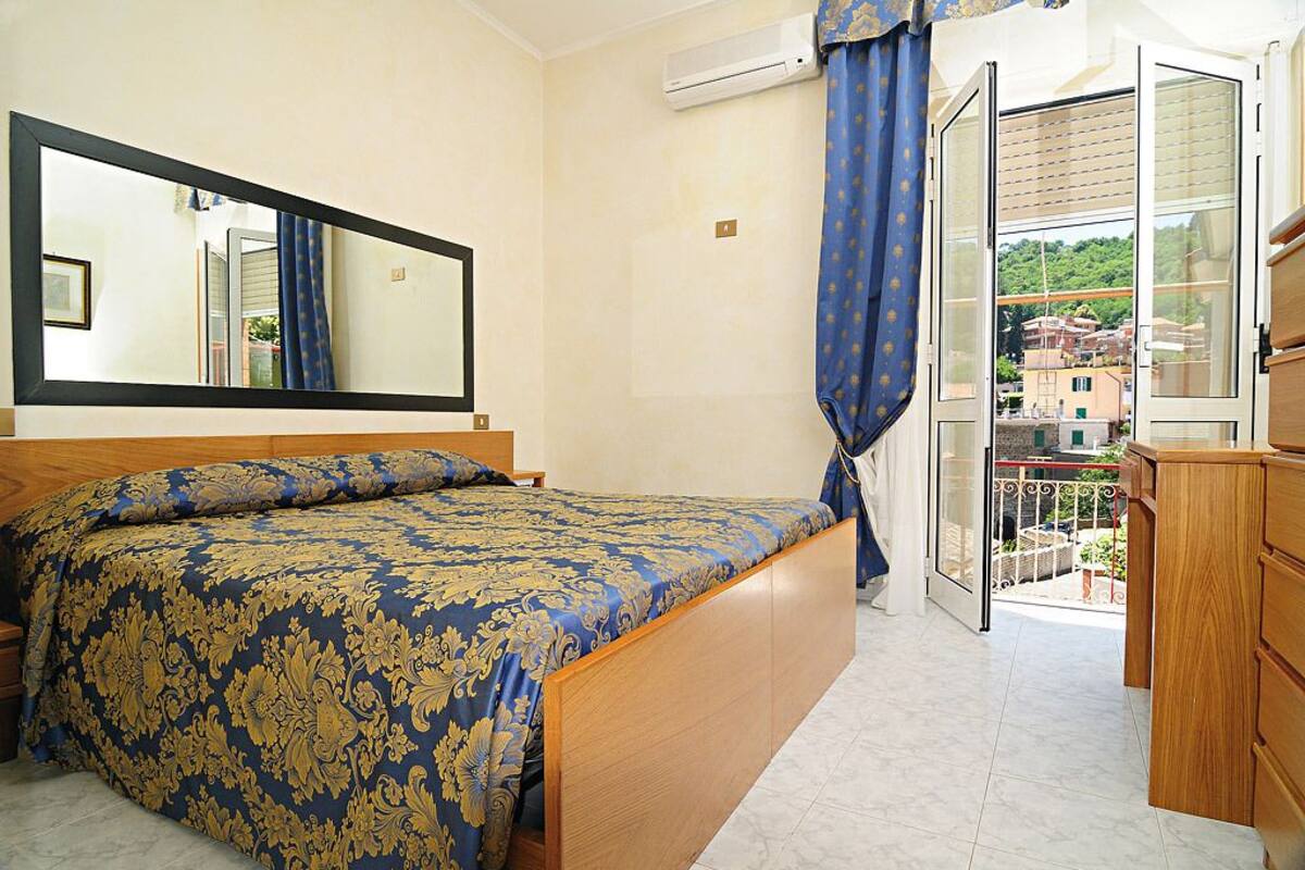 A bedroom with a bed, a large mirror, an air conditioning unit, and a balcony with a view of buildings.