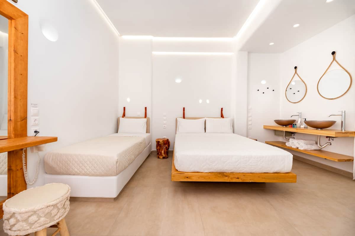 A modern hotel room with two beds, a wooden bedside table, a small stool, a wooden bench, and a bathroom with a wooden vanity and a bowl sink.