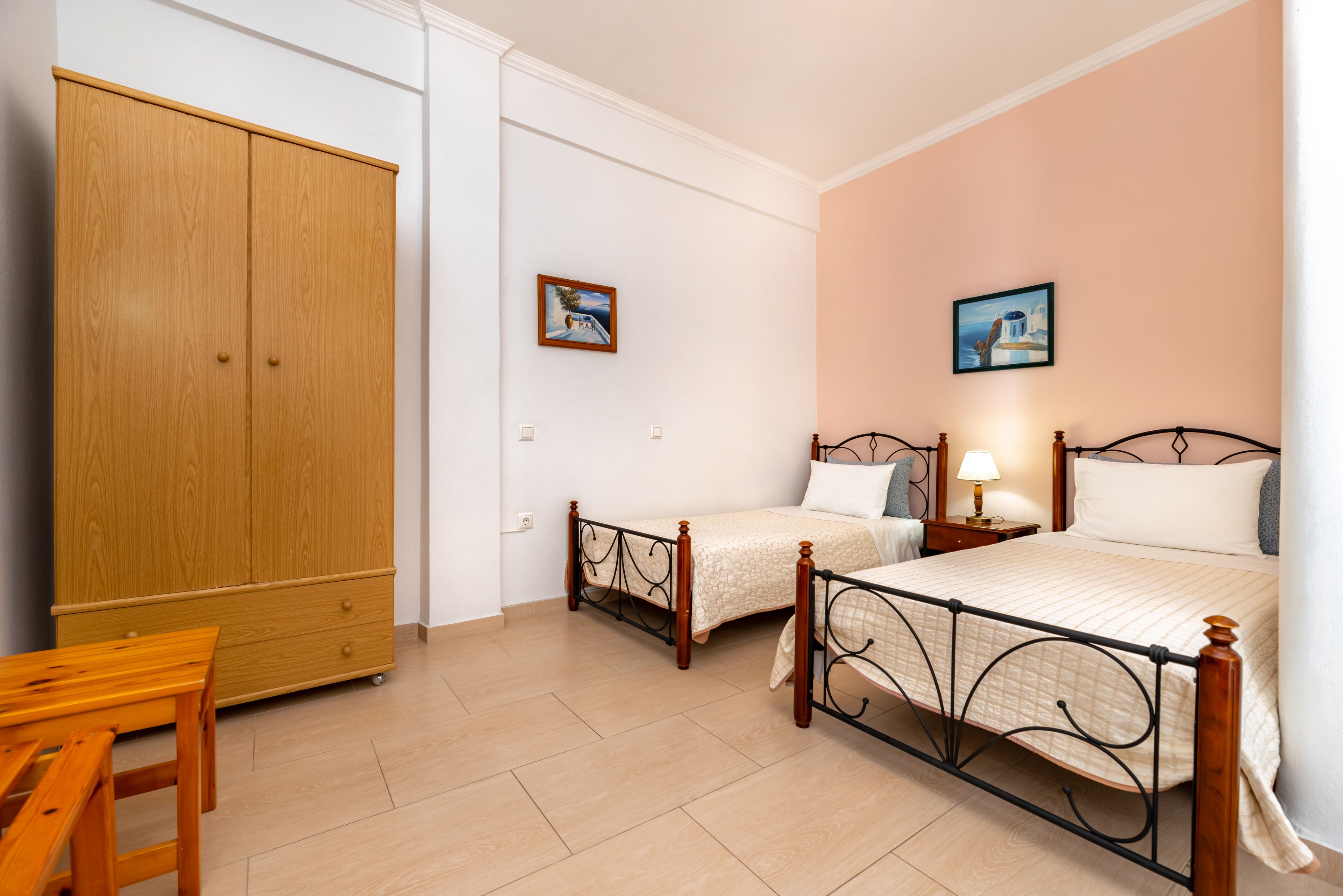 A bedroom with two single beds, a wooden wardrobe, a small table, and two wall-mounted pictures.